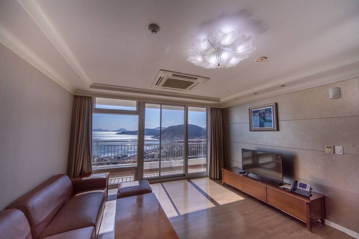 Amplia sala de estar con un ventanal que ofrece vistas al mar y a las montañas, un sofá moderno, un televisor de pantalla plana y una mesa consola de madera.