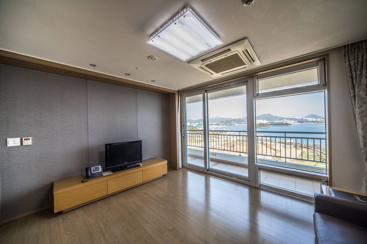 Una sala de estar moderna con un televisor de pantalla plana sobre una consola de madera, puertas de cristal correderas que dan a un balcón con vista al mar.