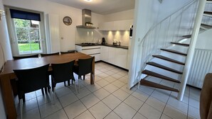 A modern kitchen with white cabinets, a dining table with chairs, a staircase, and a view of greenery outside.