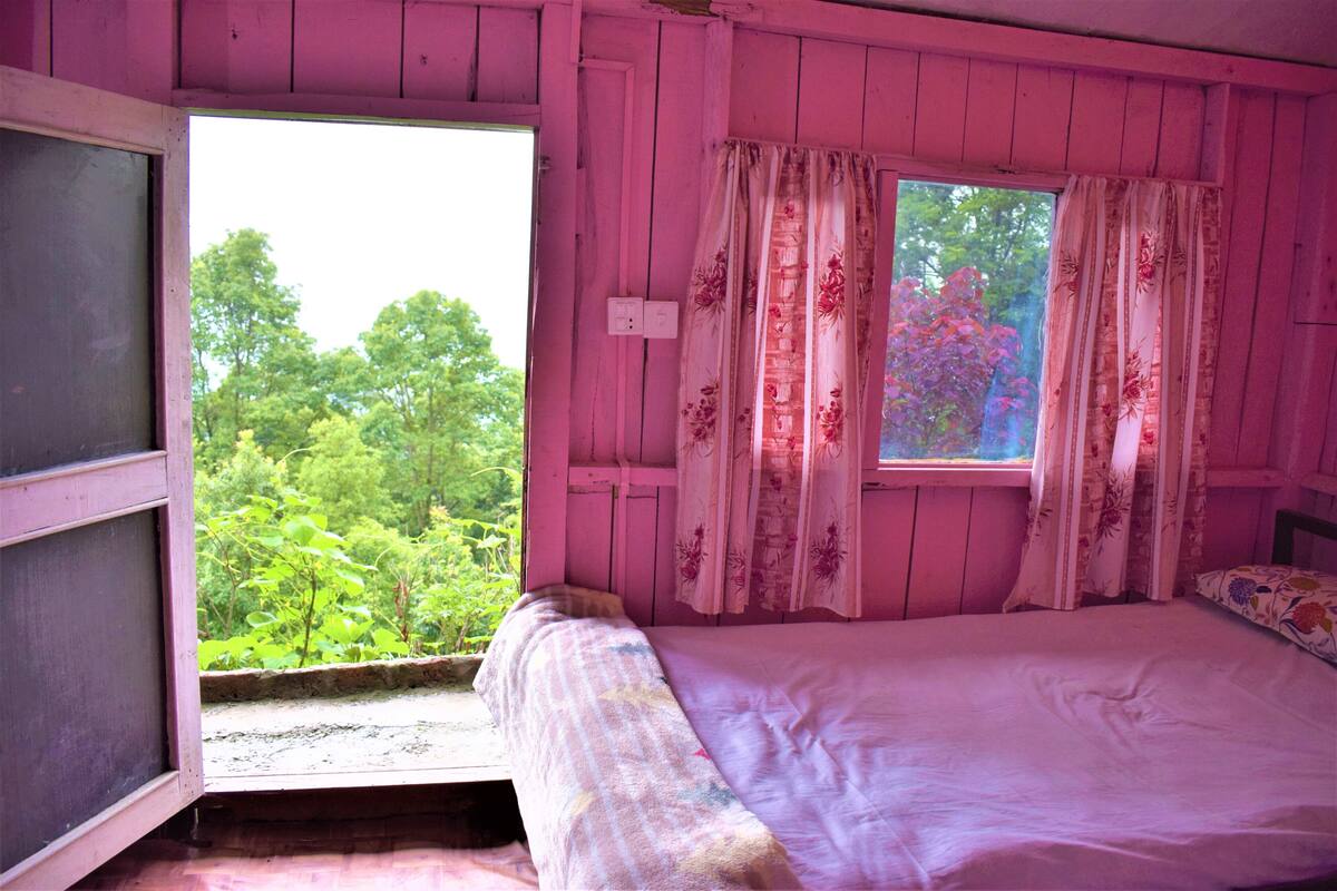 Une chambre rose avec un lit, une fenêtre avec des rideaux et une vue sur la verdure à l’extérieur.