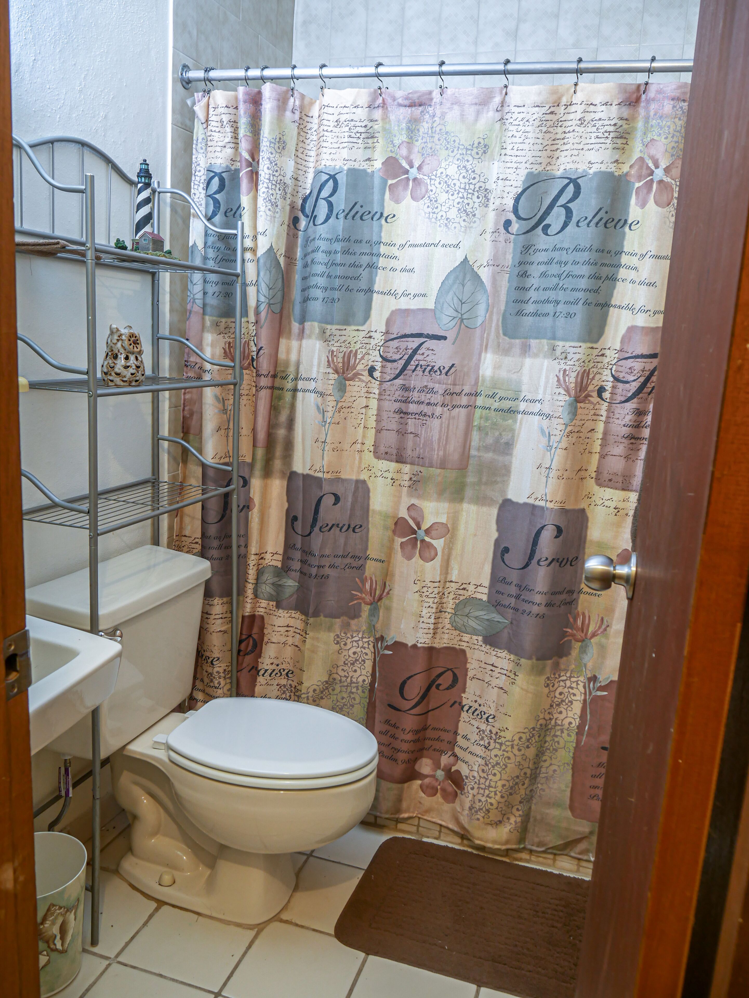 Un baño con inodoro, una estantería metálica y cortina de ducha con texto religioso.