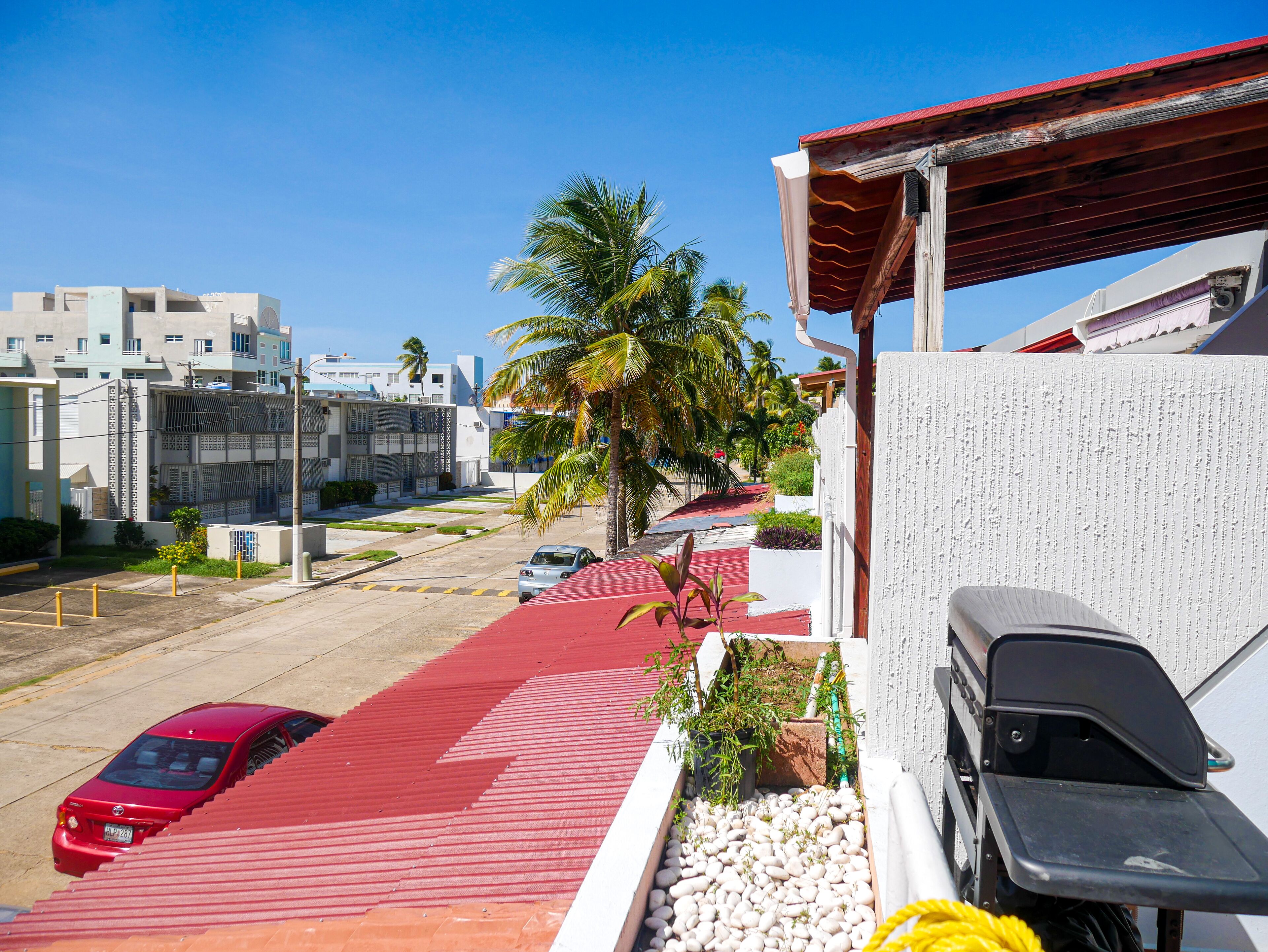 A red car parked on a red pavement, a white building with a red roof, and a palm tree in the background.