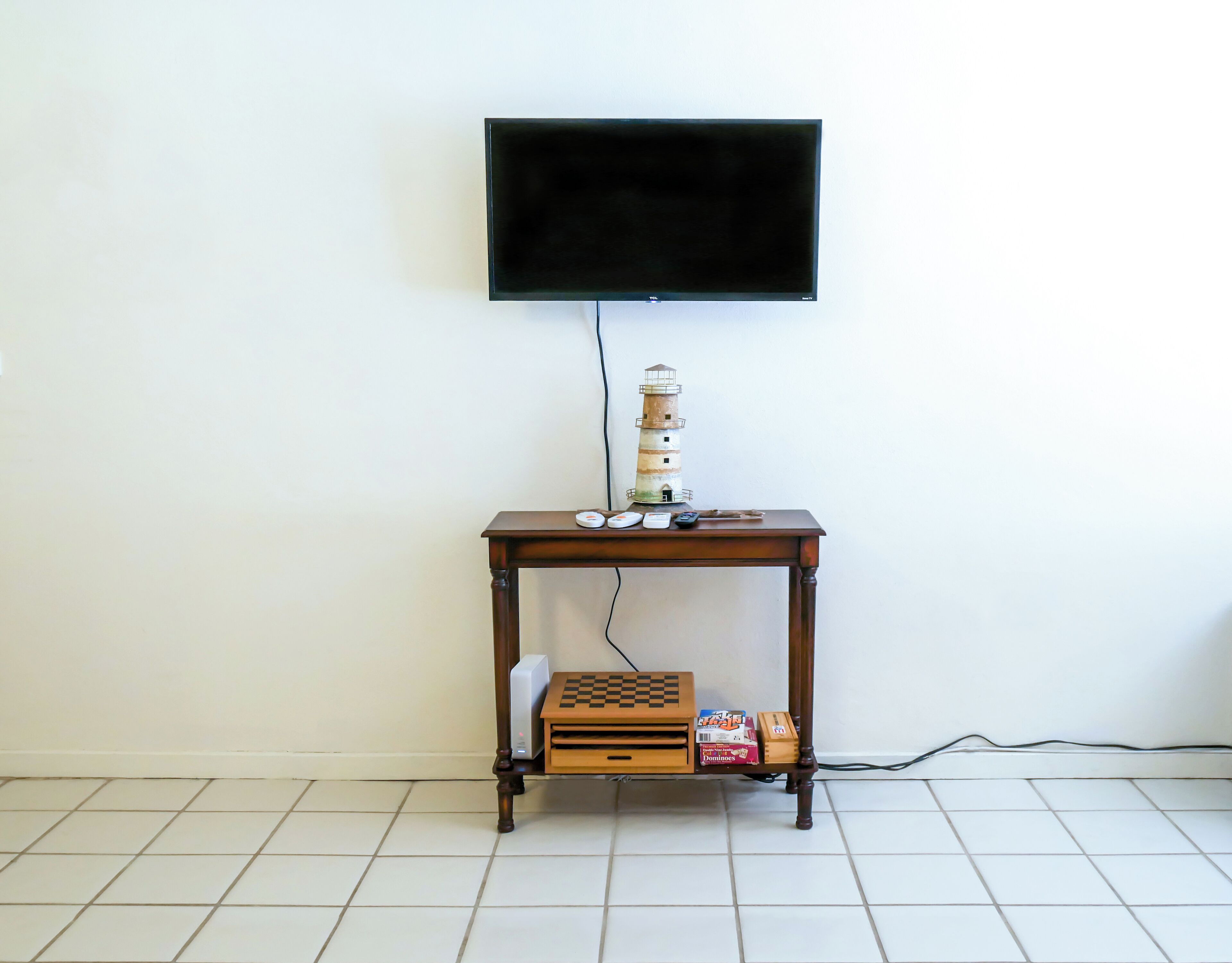 Une pièce avec un téléviseur fixé au mur, une table en bois sur laquelle se trouvent un jeu d’échecs, des dominos et une petite lampe.