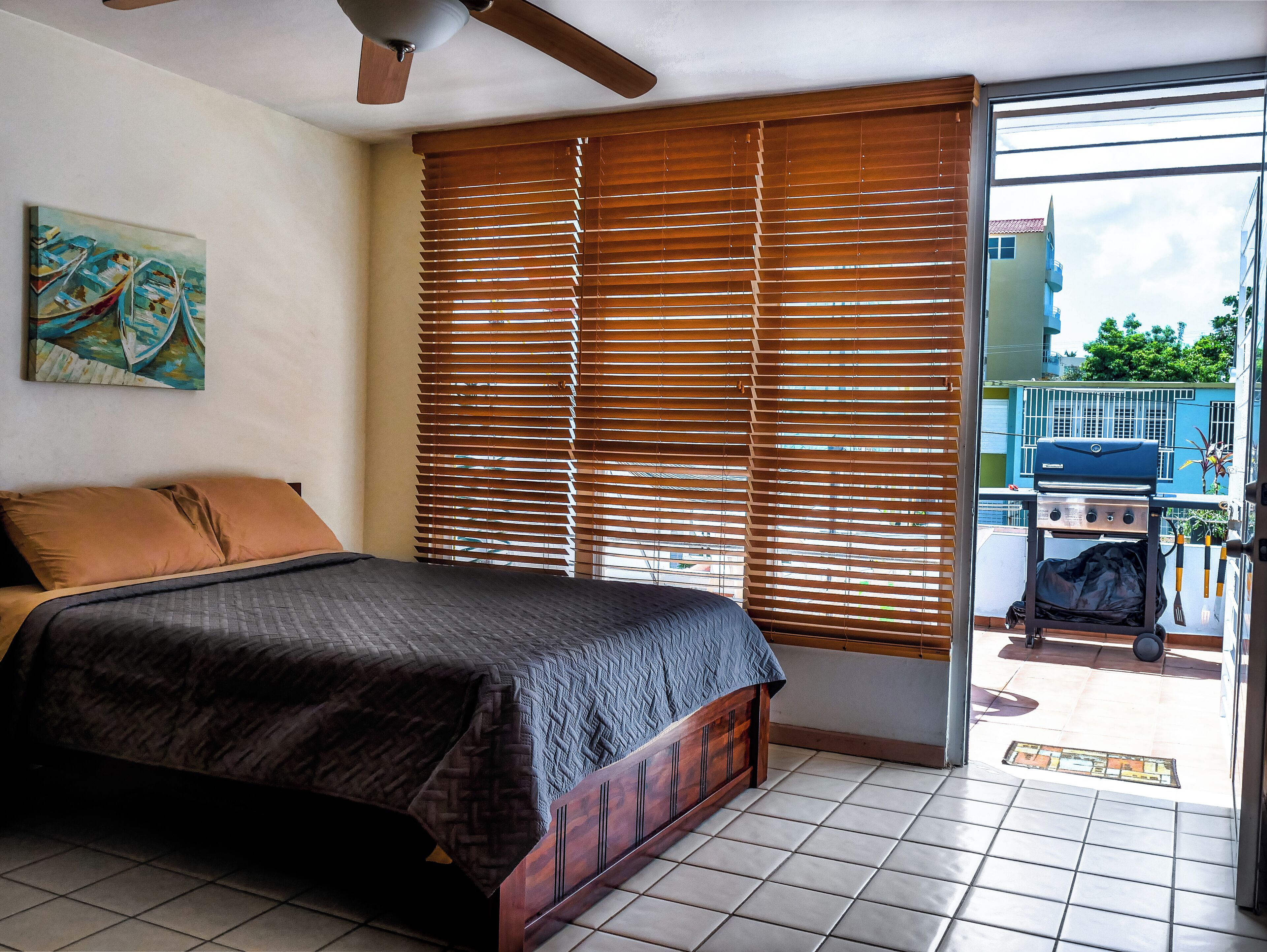 A bedroom with a bed, wooden blinds, a ceiling fan, and a view of the outside balcony.