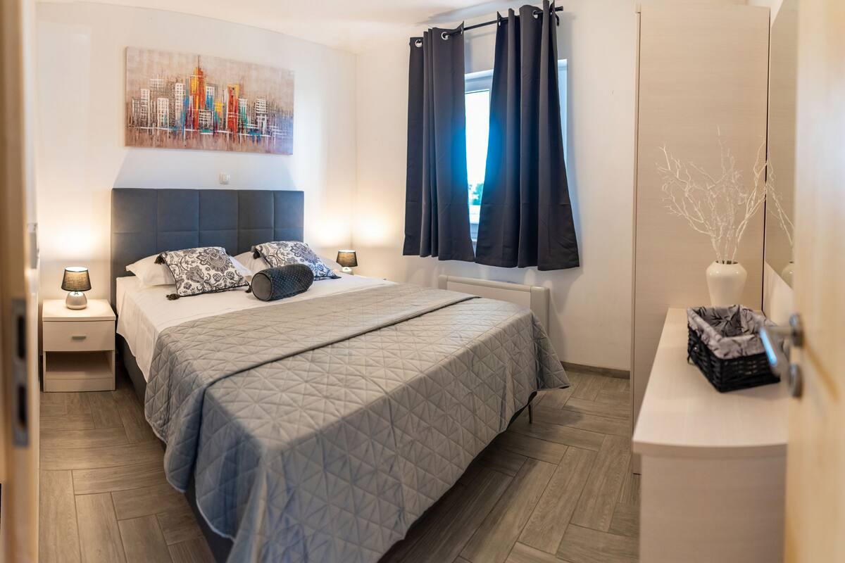 A modern bedroom with a large bed, a cityscape painting, a nightstand with a lamp, and a window with curtains.