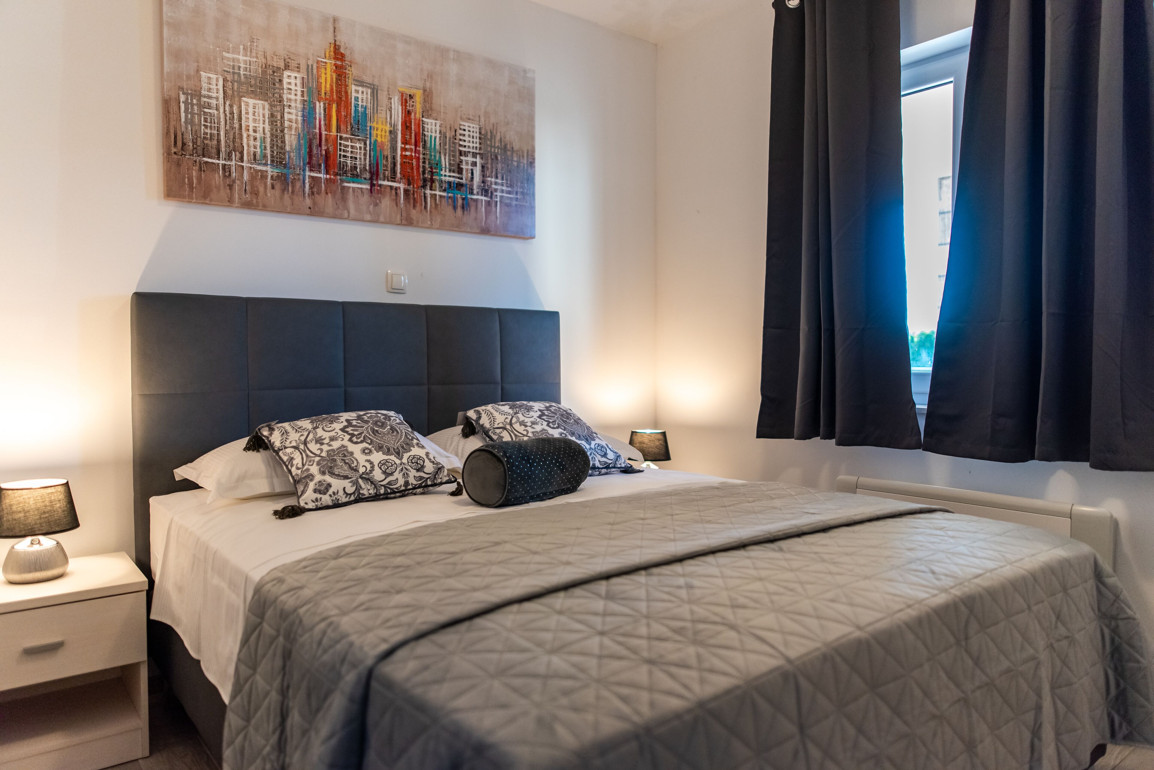 Una cama bien hecha con colcha acolchada, dos almohadas y una lámpara de noche sobre una mesita.