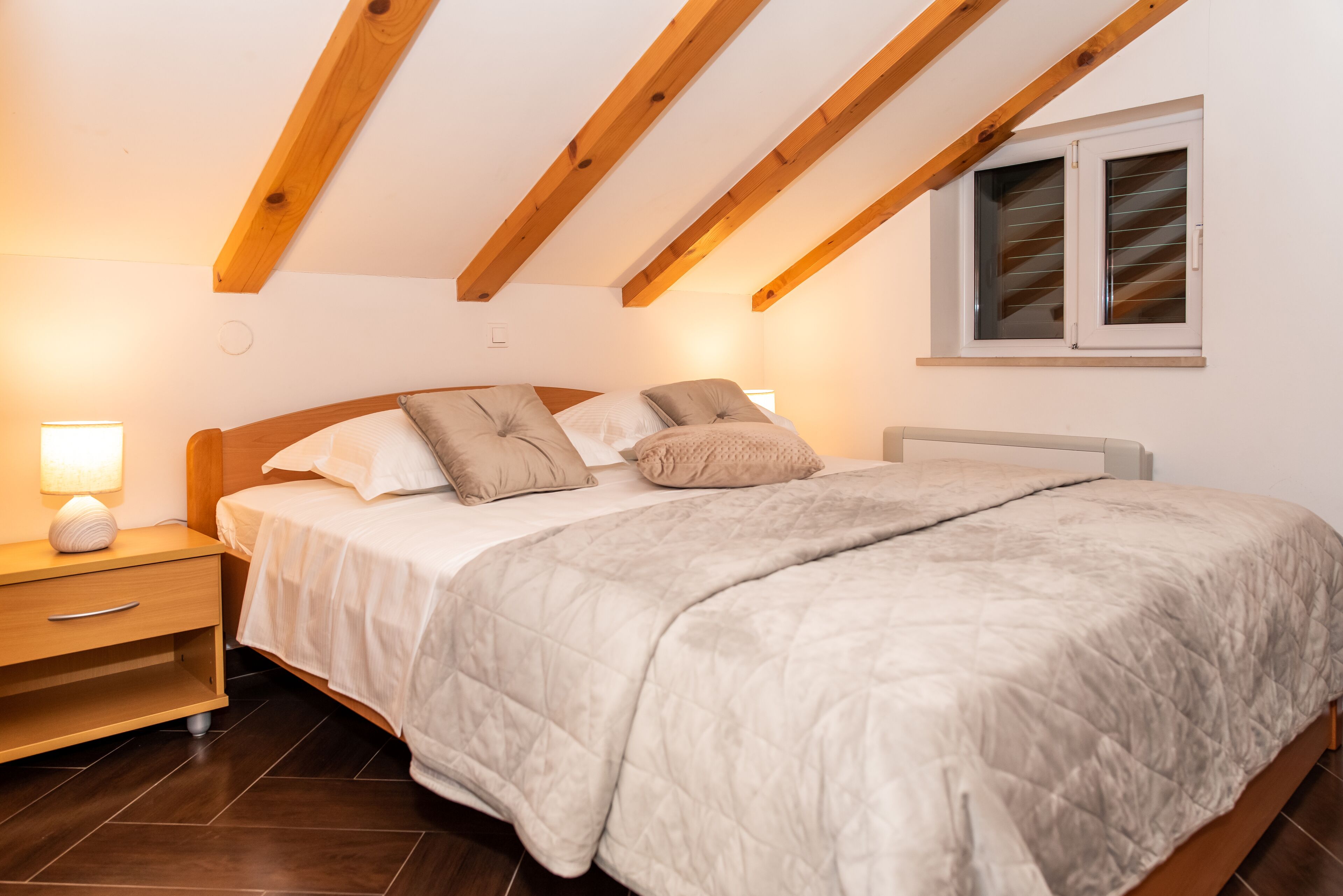 Een slaapkamer met een bed, houten hoofdbord, nachtkastje, lamp en een raam met jaloezieën.