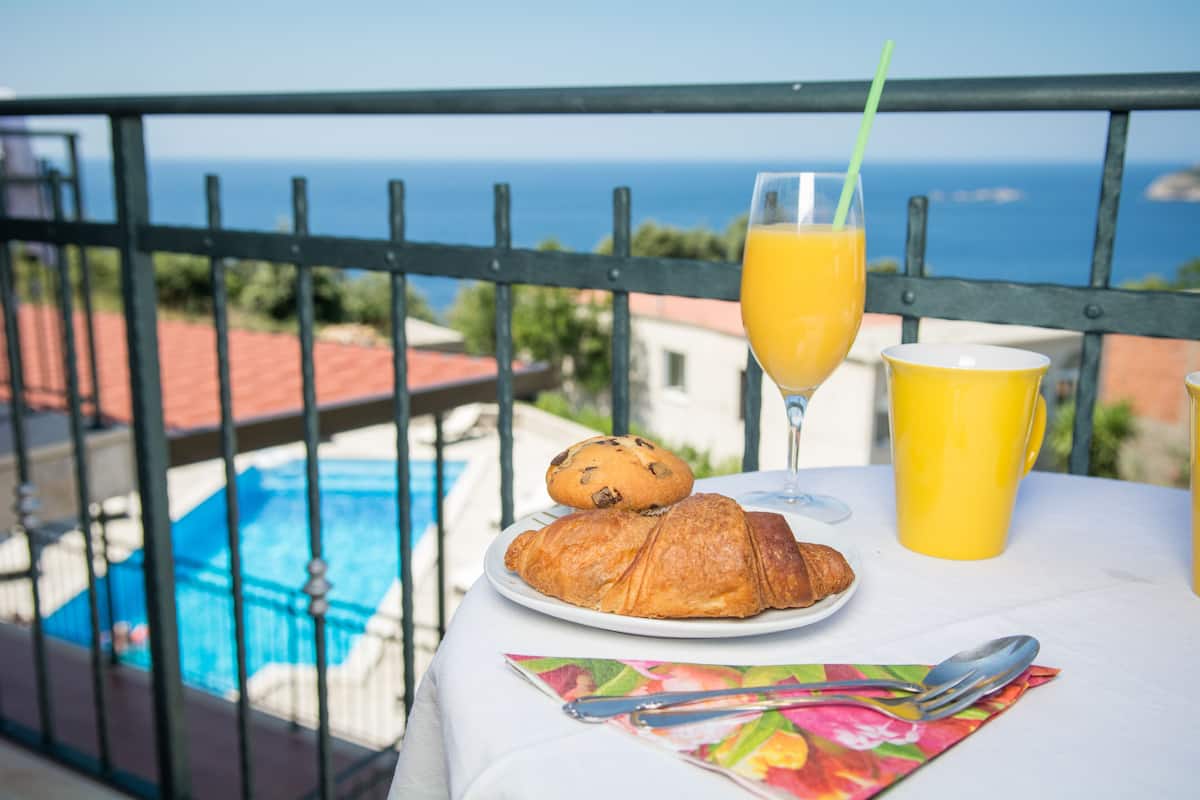 A table with a croissant, a glass of orange juice, and a yellow cup on a balcony overlooking a pool and the sea.