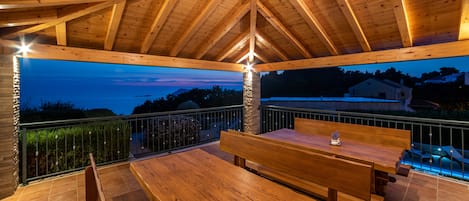 Eine hölzerne Außenterrasse mit Tisch und Bänken, mit Blick auf einen Swimmingpool und ein Haus im Hintergrund.