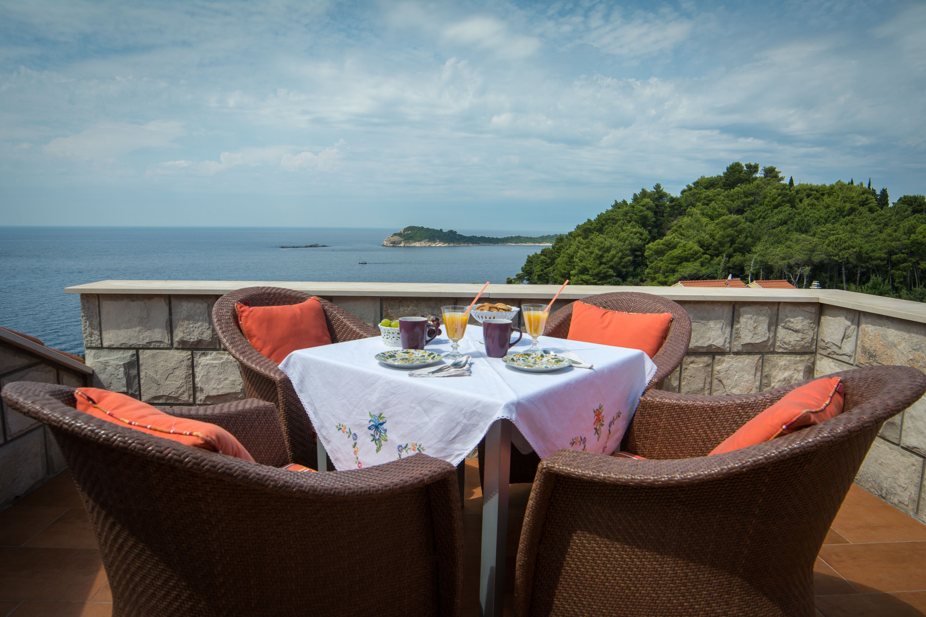 Une table à manger recouverte d’une nappe blanche, entourée de chaises en osier agrémentées de coussins orange, est installée sur une terrasse dotée d’un mur en pierre et offrant une vue sur la mer et une île lointaine.