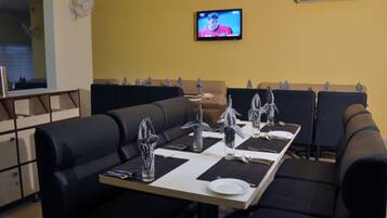 A restaurant with a long table set for dining, black cushioned booths, a television on the wall, and an air conditioning unit.