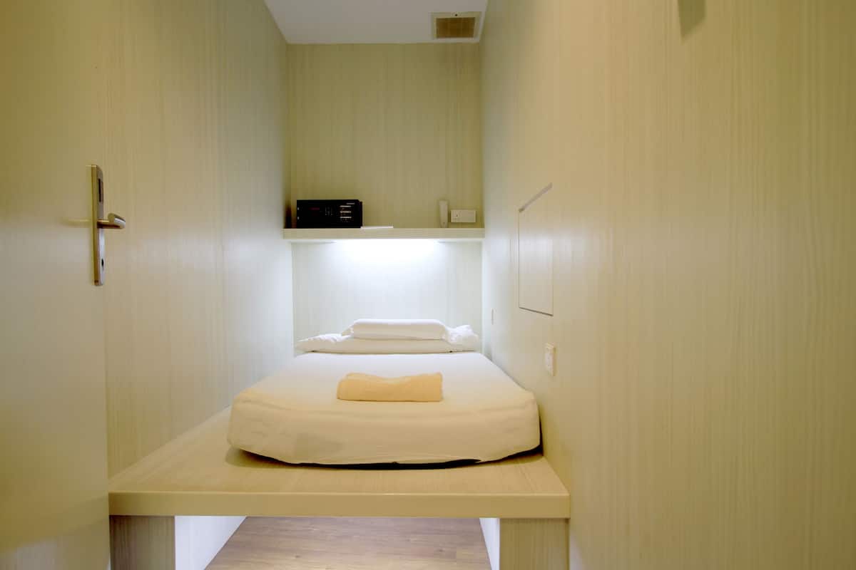 A small, single-bed room with a wooden bed frame, a bedside table, a wall-mounted shelf with a black device, and a white wall switch.