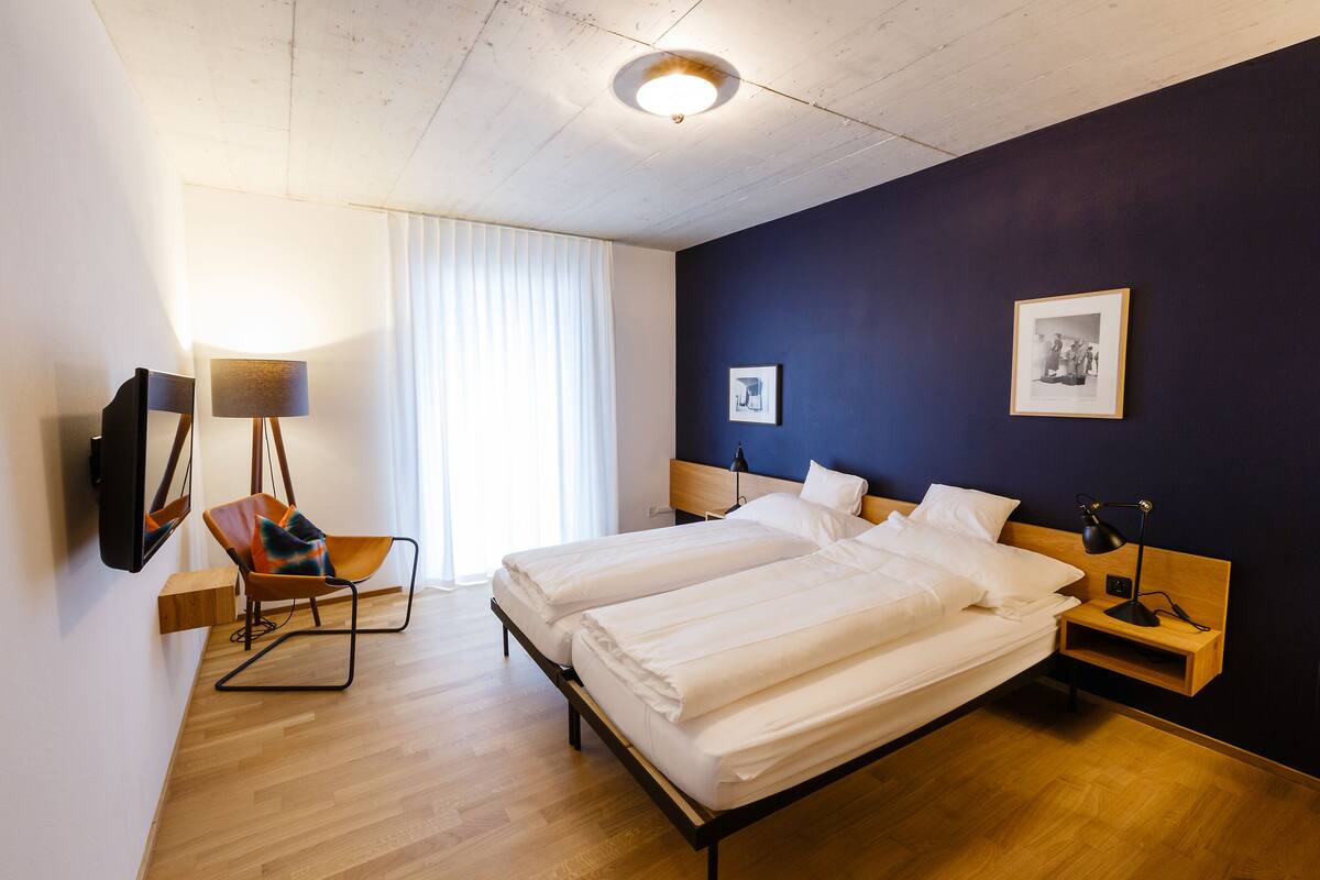 Habitación de hotel con dos camas, una silla, un escritorio con lámpara, un cuadro en la pared y una ventana con cortinas.