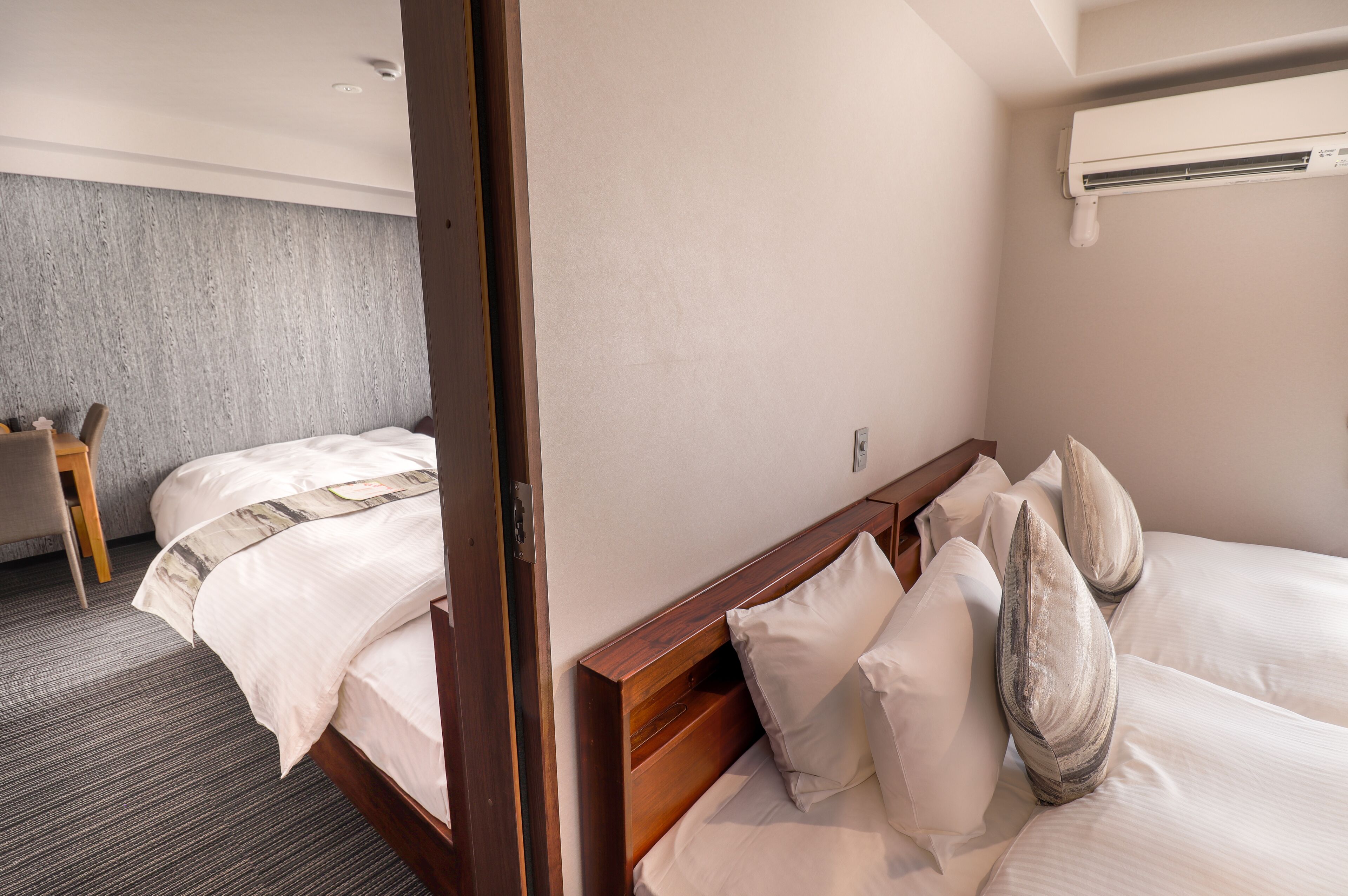 Une chambre d’hôtel avec une tête de lit en bois, un lit recouvert de draps blancs et agrémenté de coussins décoratifs, un mur gris à texture, et une petite table en bois.