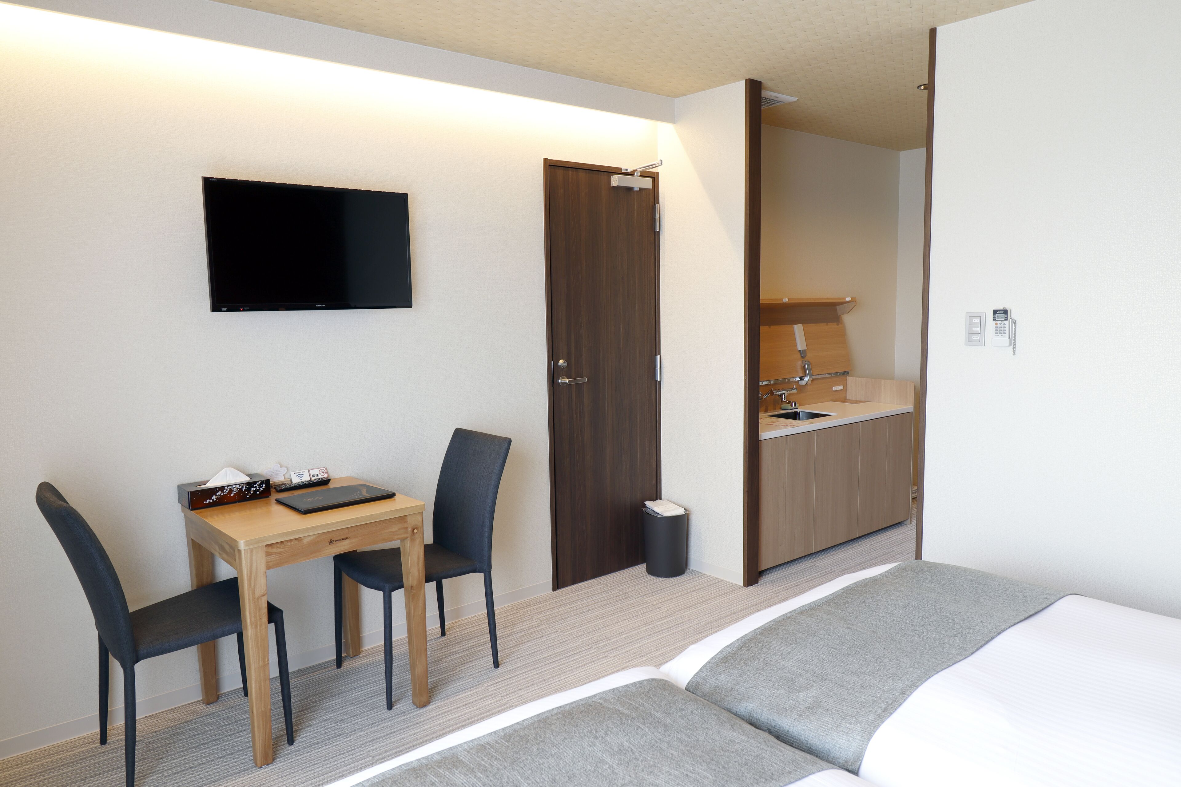 Une chambre d’hôtel comprenant un lit, un bureau avec une chaise, une télévision, une porte en bois et une petite kitchenette.