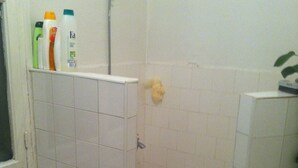A small bathroom with white tiles, a shower, and a toilet. There are toiletries on the wall.