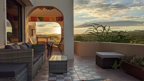 A balcony with wicker furniture, a table, and chairs, overlooking a mountainous landscape at sunset.