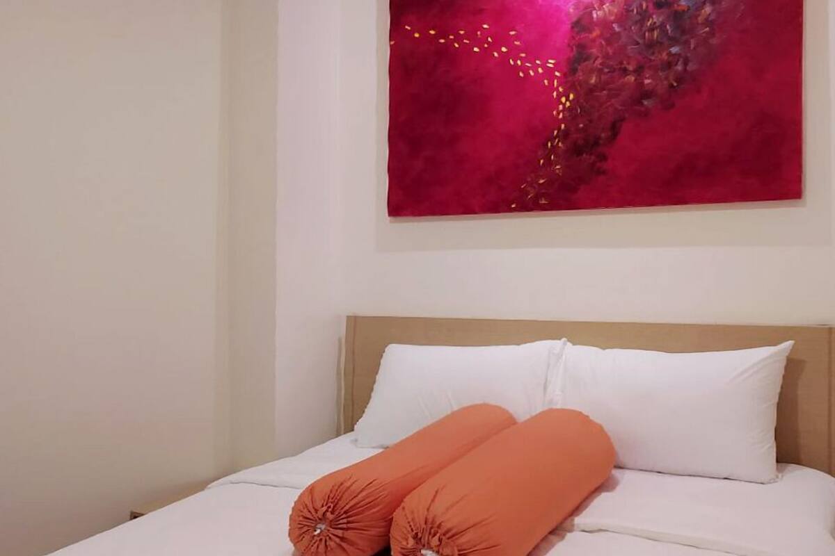 A neatly made bed with white bedding and two orange pillows, a red abstract painting on the wall, and a wooden headboard.