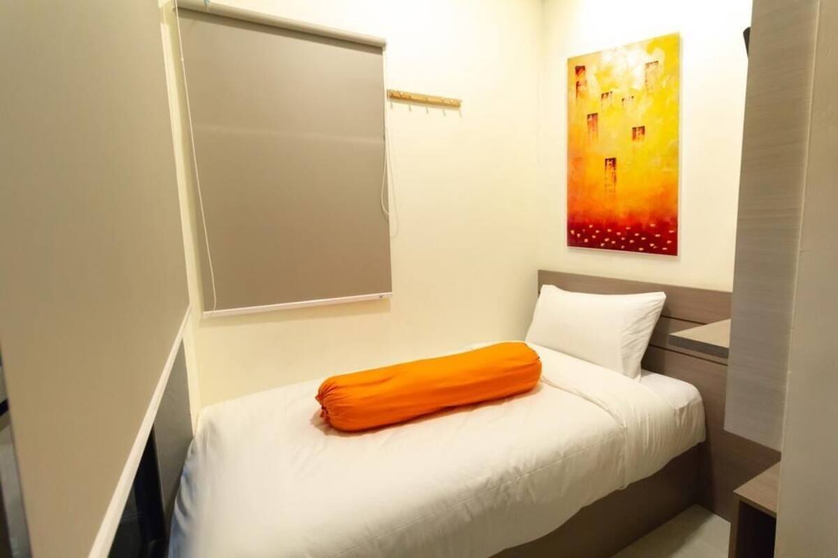 A single bed with white bedding and an orange pillow, a wall-mounted shelf, and a painting on the wall.