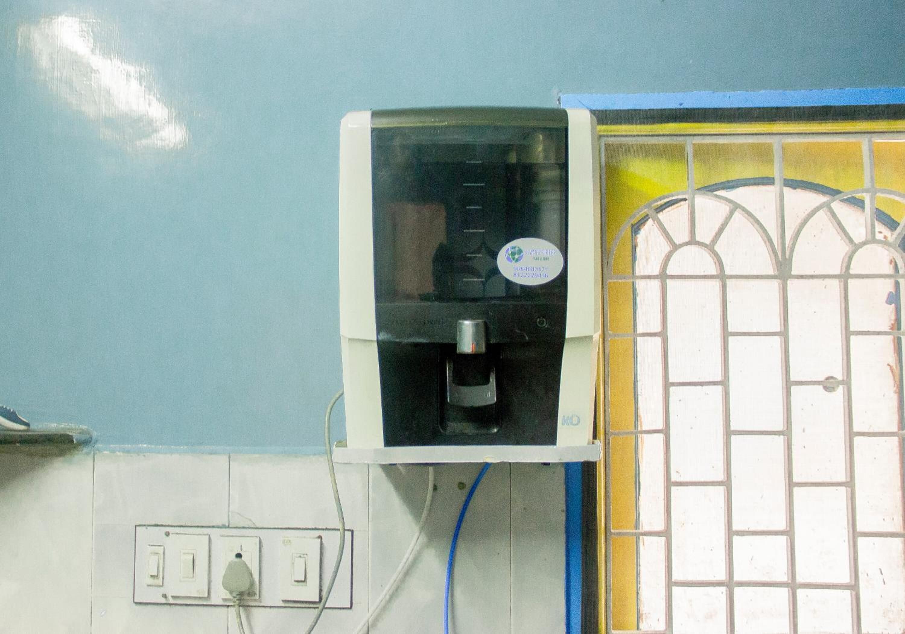 Un dispensador de agua fijado en una pared, junto a una ventana y una puerta con celosía metálica.