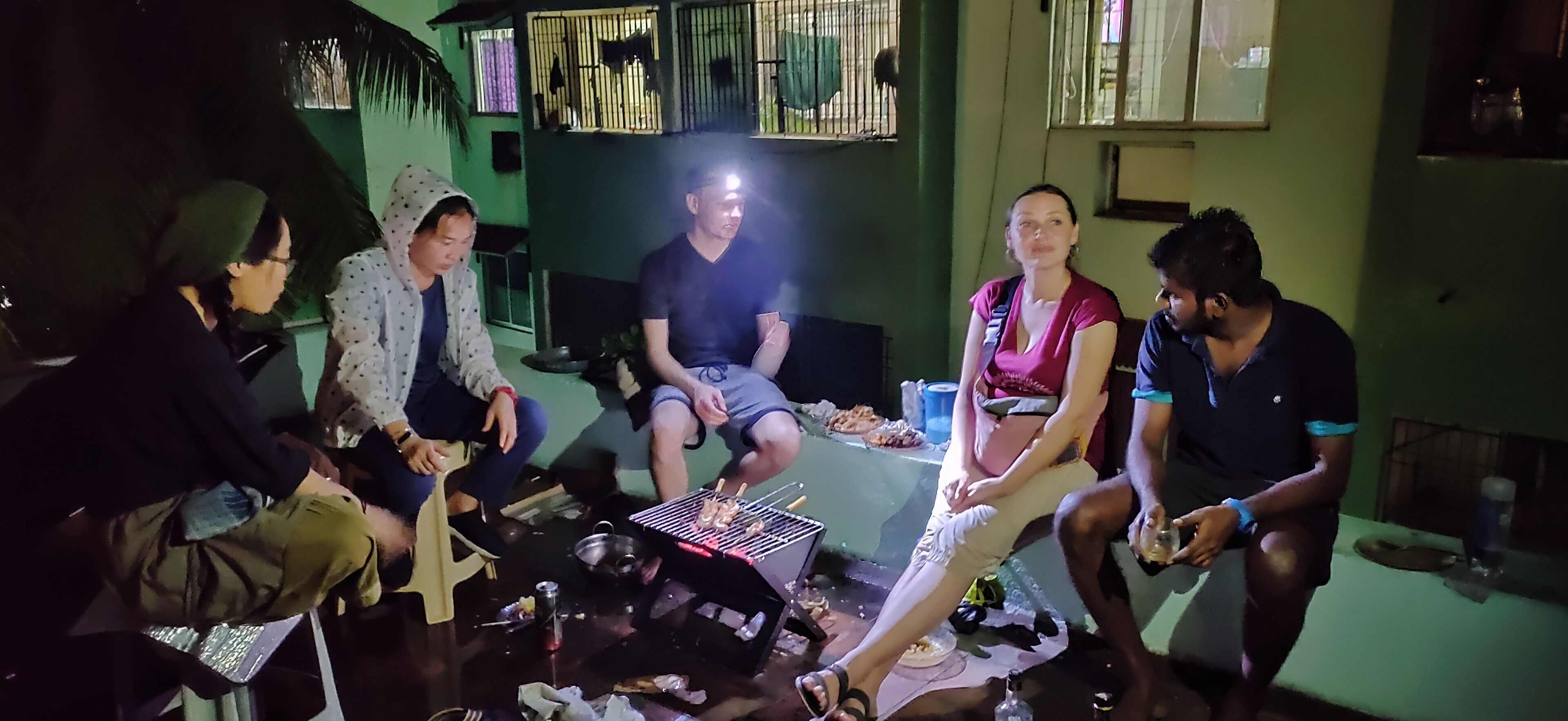 Eine Gruppe von Personen sitzt an einem Tisch mit einem Grill in einem Raum mit grünen Wänden und einem Fenster mit Gitterstäben.