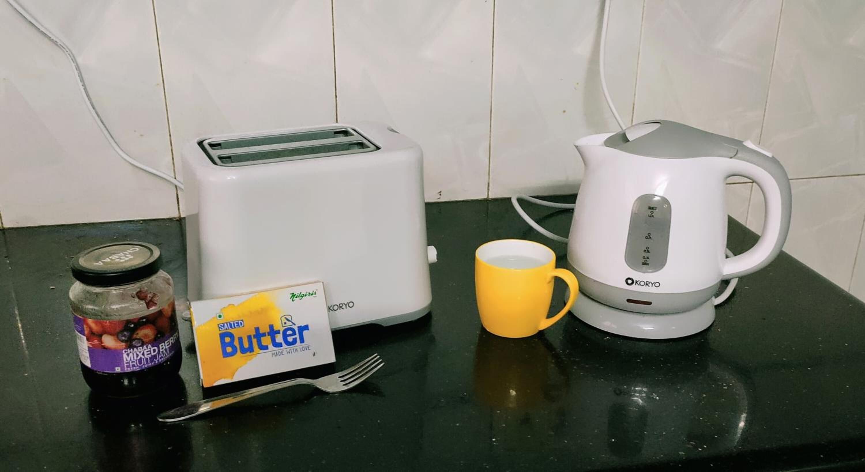 Eine Küchenarbeitsfläche mit einem Toaster, einem Wasserkocher, einem Glas gemischter Fruchtkonfitüre, einer Butterportion, einer gelben Tasse und einer Gabel.
