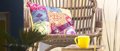 A wicker chair with a colorful quilted pillow and a yellow cup on a small wooden table.