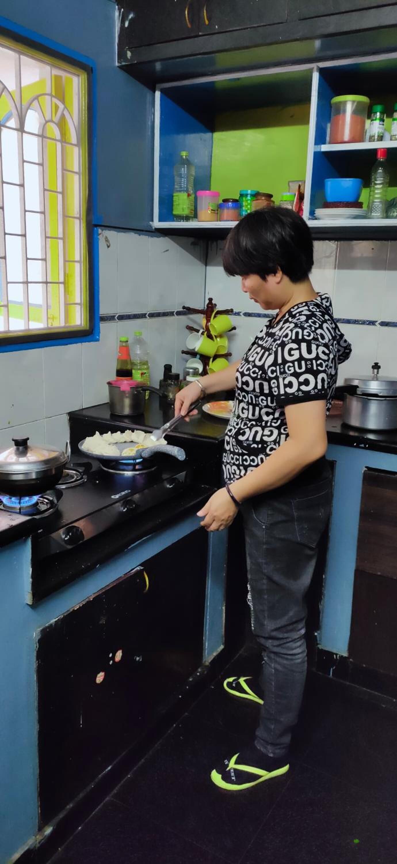 A person cooking on a stove in a kitchen with blue walls and a window with a yellow frame.