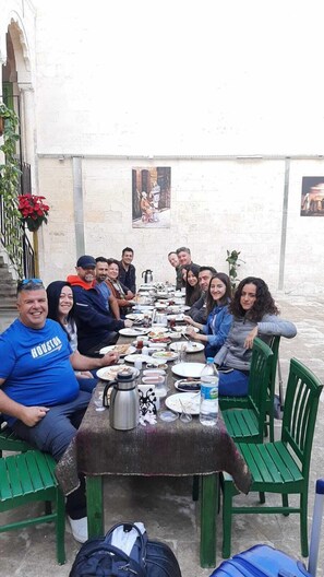 A group of people seated around a table with various dishes and drinks, in an outdoor setting with a white wall and a green bench. - Hanehan Butik Otel (Sanliurfa)