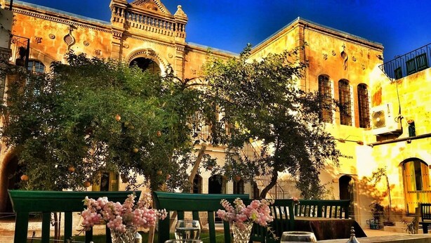 An outdoor dining area with a table set for two, surrounded by green benches, in front of a historic building with arches and a clock tower. - Hanehan Butik Otel (Sanliurfa)