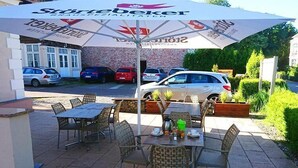Un espacio al aire libre con sillas y mesas de mimbre, un paraguas grande y vistas a coches aparcados y un edificio al fondo.