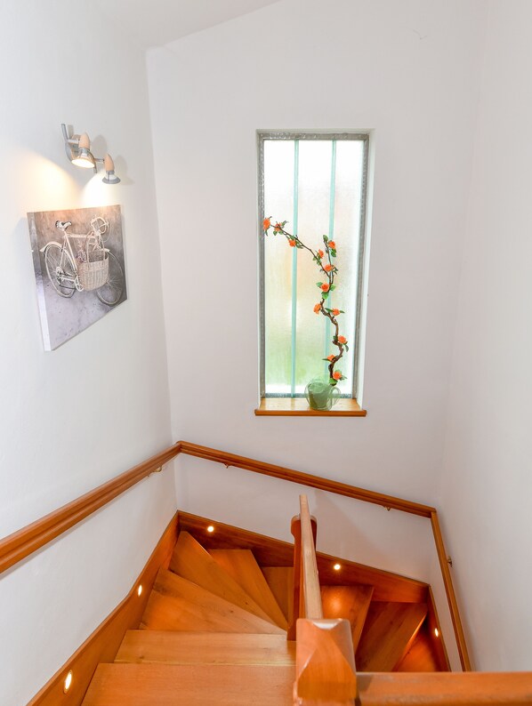 Una escalera de madera con barandilla, un cuadro colgado en la pared de una bicicleta y una ventana con una planta.