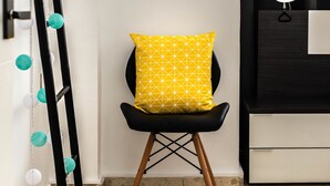 Een stoel met een geel kussen, een zwart-wit lamp en een spiegel aan de muur.