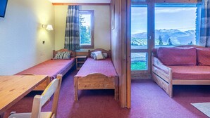 A cozy room with two wooden beds, a wooden table, and a sofa. There is a window with a view of mountains and trees. - Divisible studio for 4 in Valmorel near slopes, shops and lifts (Valmorel)