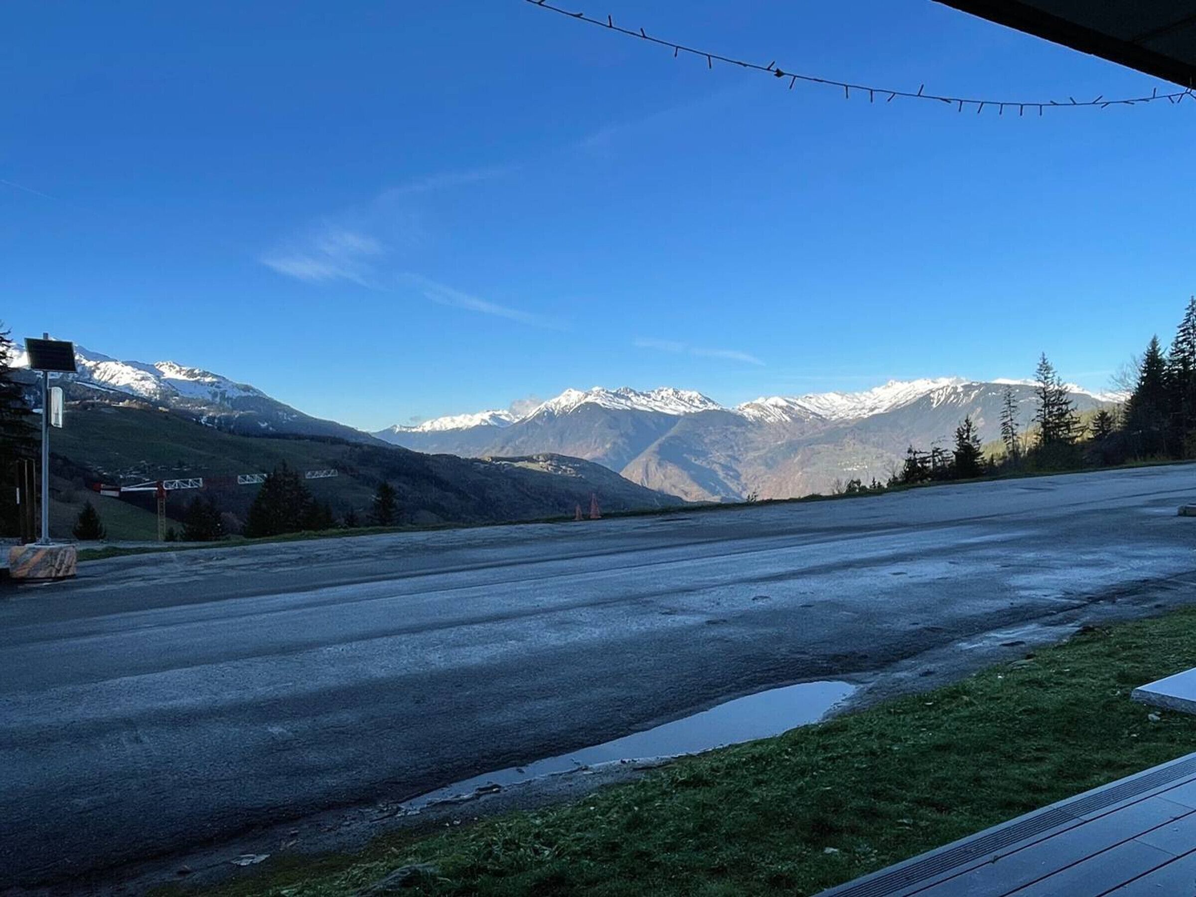 Une route bordée de montagnes enneigées en arrière-plan, un ciel dégagé et une zone herbeuse avec un bâtiment sur la gauche.