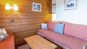 A cozy room with wooden panel walls, a pink sofa with blue cushions, a wooden table, and two framed pictures on the wall. - Divisible studio for 4 in Valmorel near slopes, shops and lifts (Valmorel)