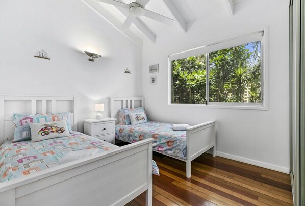 A bedroom with two single beds, white walls, a large window with a view of greenery, and a ceiling fan.