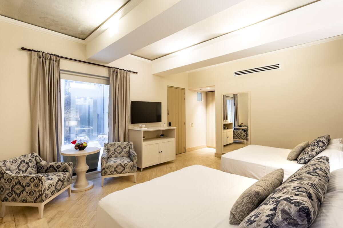 Une chambre d’hôtel avec deux lits, une télévision, une petite table avec des fruits, des chaises à motifs et une grande fenêtre avec des rideaux.