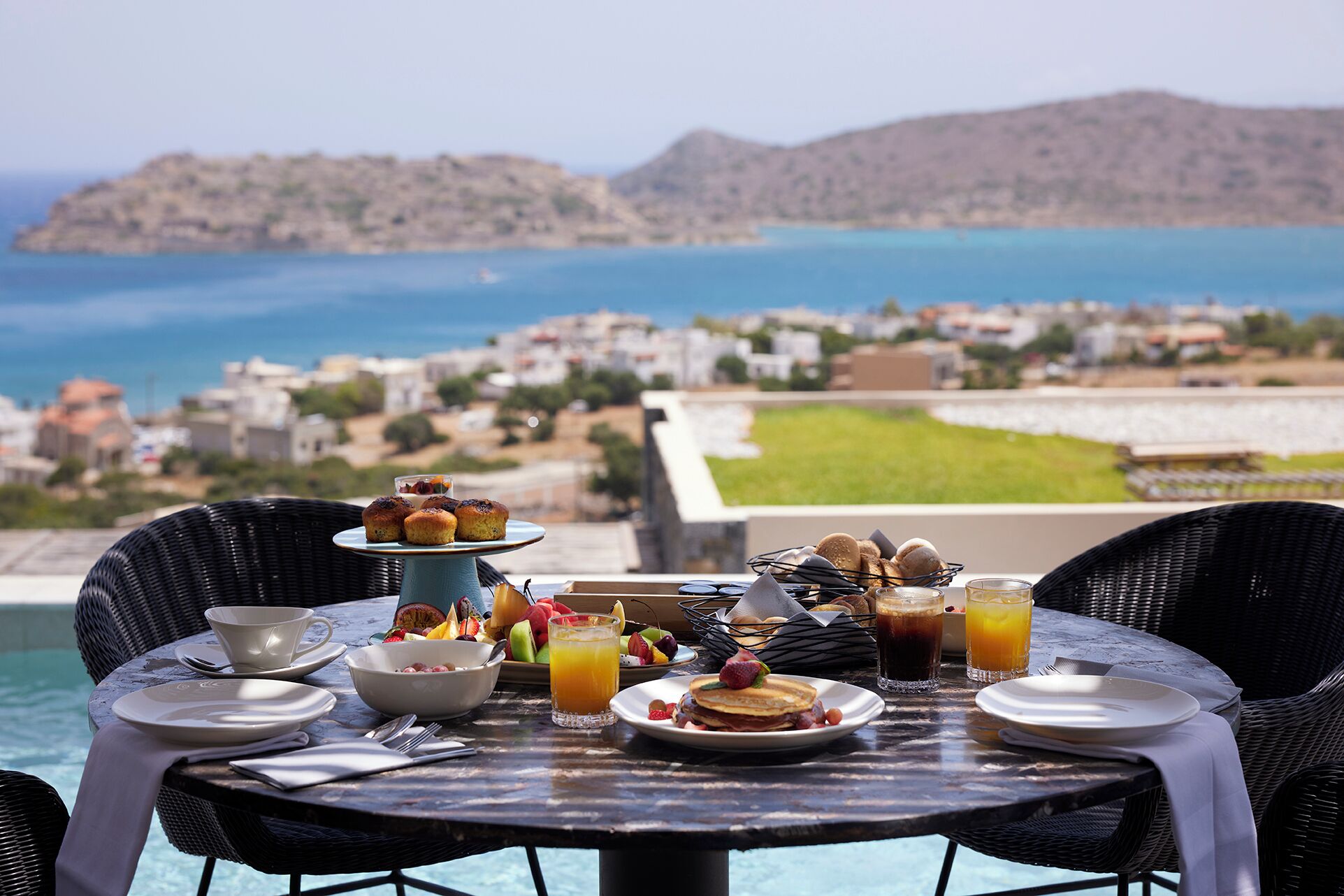 Una tavola imbandita per la colazione con vista su una cittadina costiera, con piscina e montagne sullo sfondo.