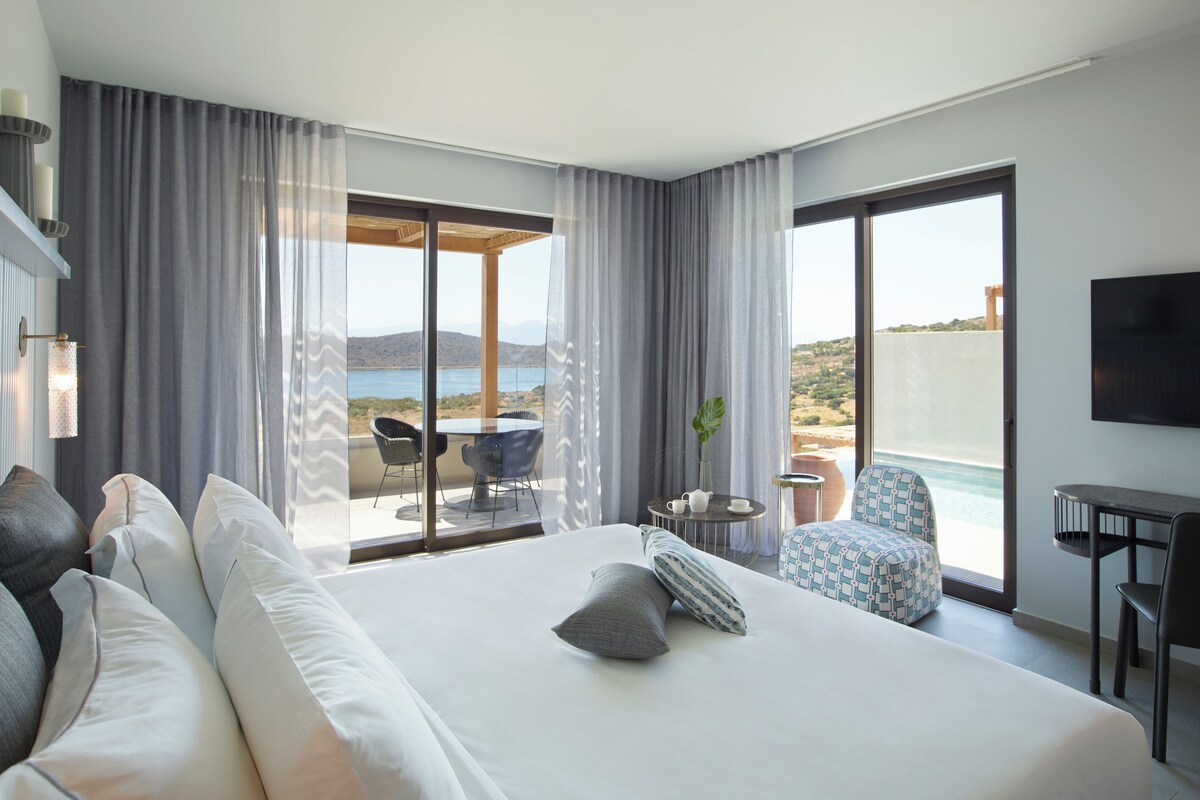 A modern hotel room with a large bed, a balcony with a table and chairs, and a view of the sea.