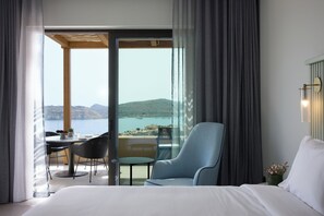 A hotel room with a bed, a chair, a table, and a view of the sea and mountains. - Cayo Exclusive Resort & Spa (Agios Nikolaos)