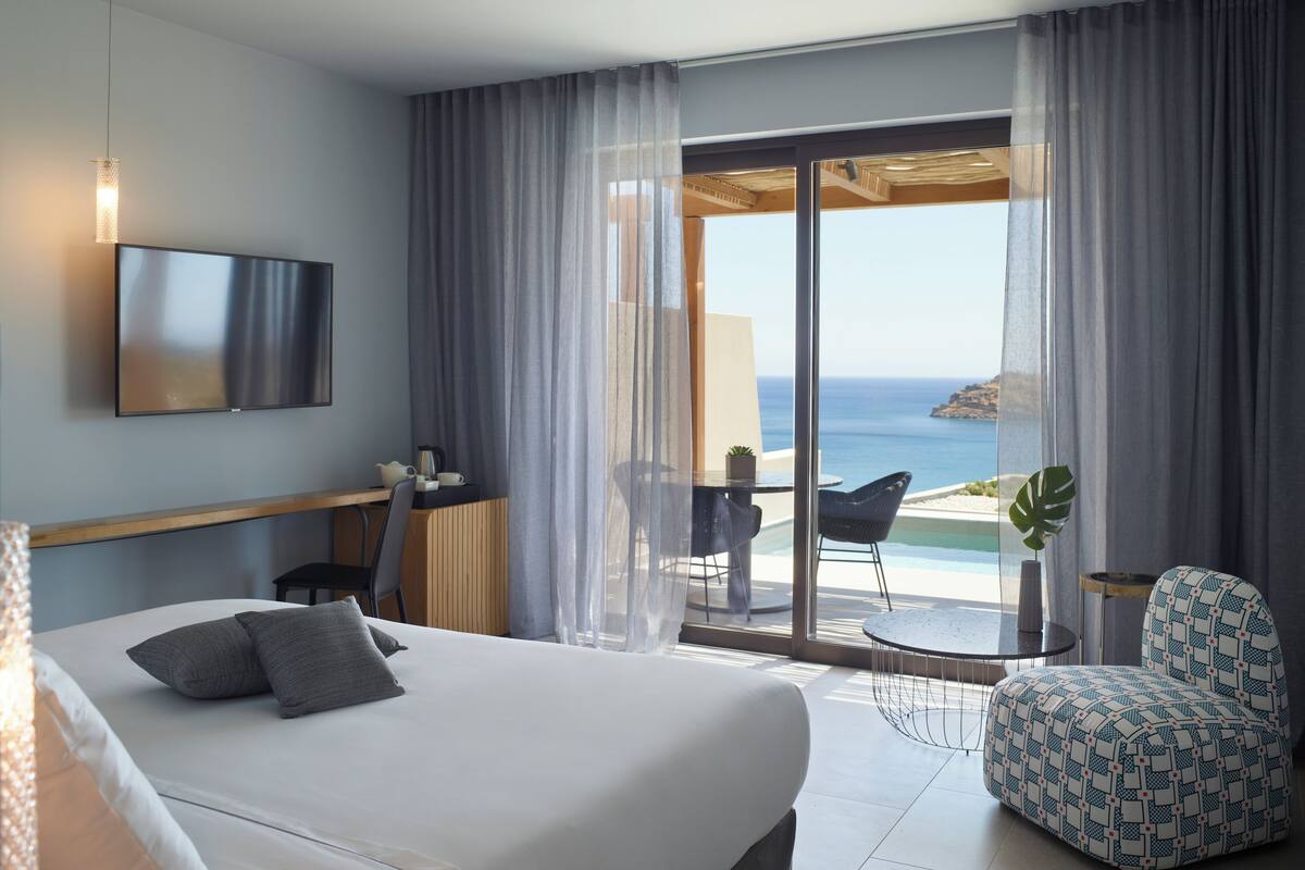 A hotel room with a bed, a TV, a desk with a chair, a small table with a chair, and a view of the sea.