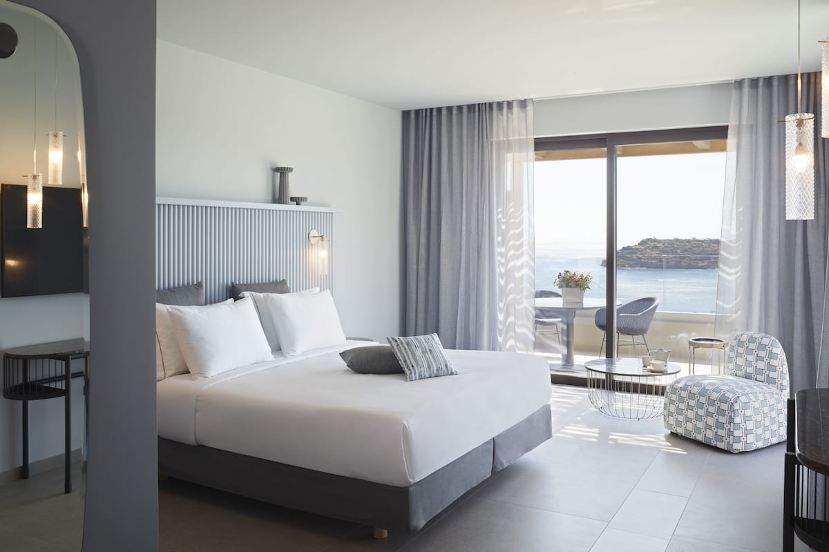 A modern hotel room with a large bed, a desk, a chair, and a view of the sea.