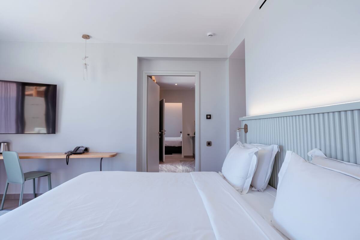A modern hotel room with a large bed, a desk with a chair, and a view into another room.