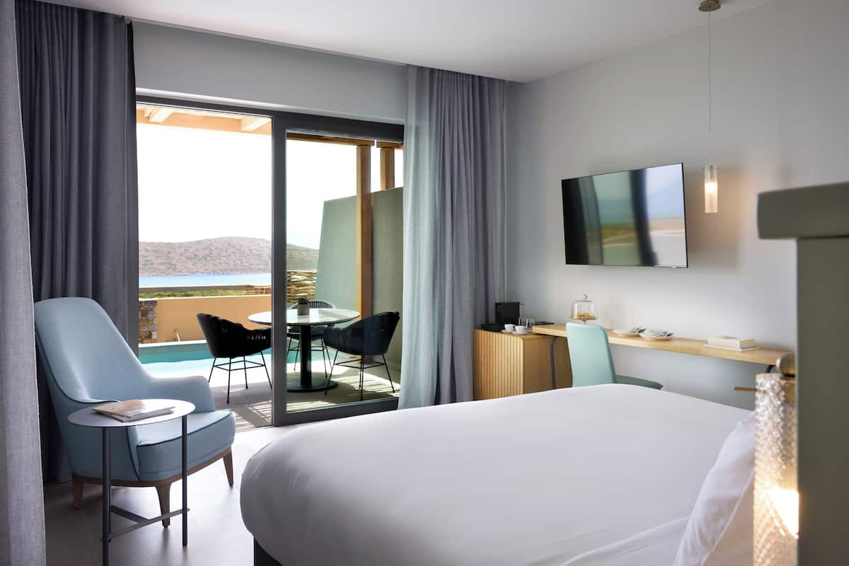 A hotel room with a bed, a desk, a chair, a TV, and a view of a pool and mountains.