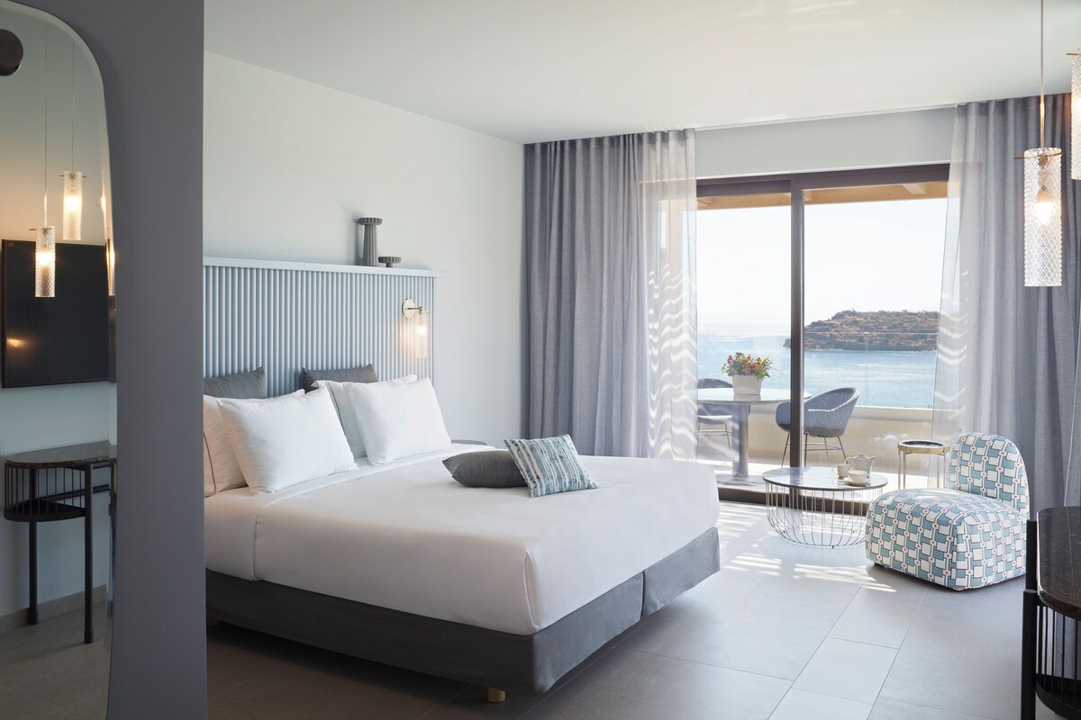 A modern hotel room with a large bed, a desk, and a view of the sea.