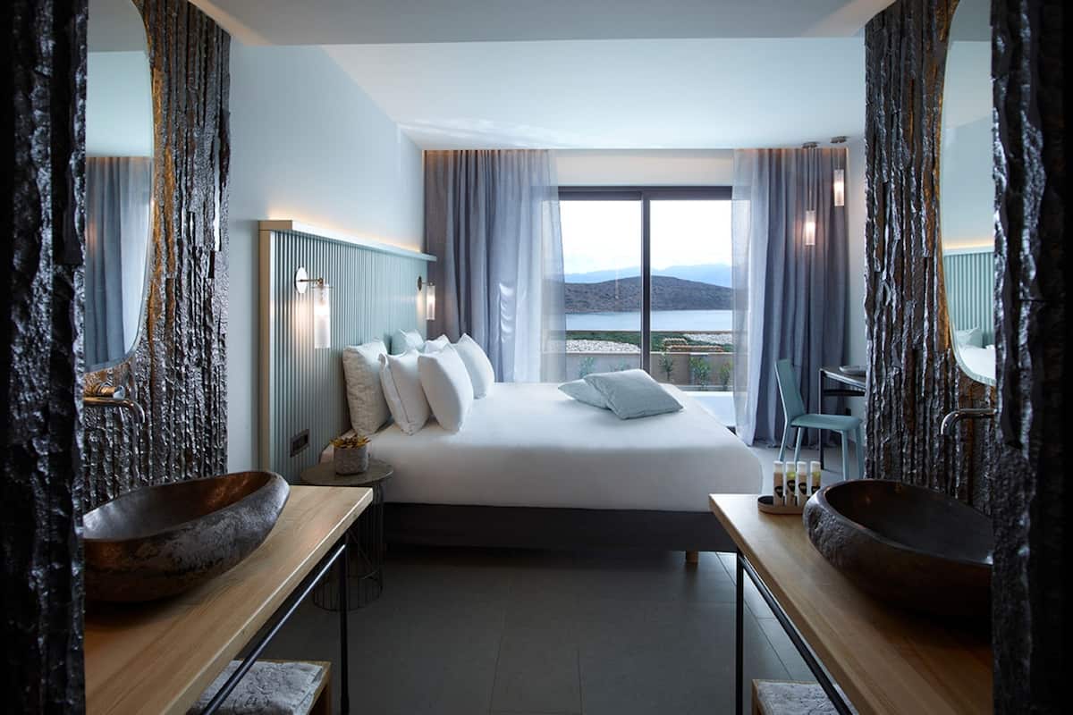 A modern hotel room with a large bed, two side tables with bowls, a view of the sea, and a stone-textured wall.
