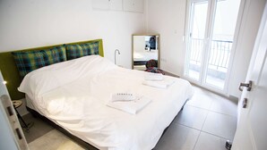 A neatly made bed with white linens and two folded towels with text, a green headboard, a bedside table with a lamp, and a window with a view of the outside. - Zennova 9 City Center Pearl (Thessaloniki)