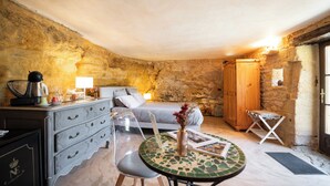 A rustic room with a bed, wooden wardrobe, clear chairs, a small round table with a vase, and a stone wall. - La Suite Troglodyte (Vitrac)