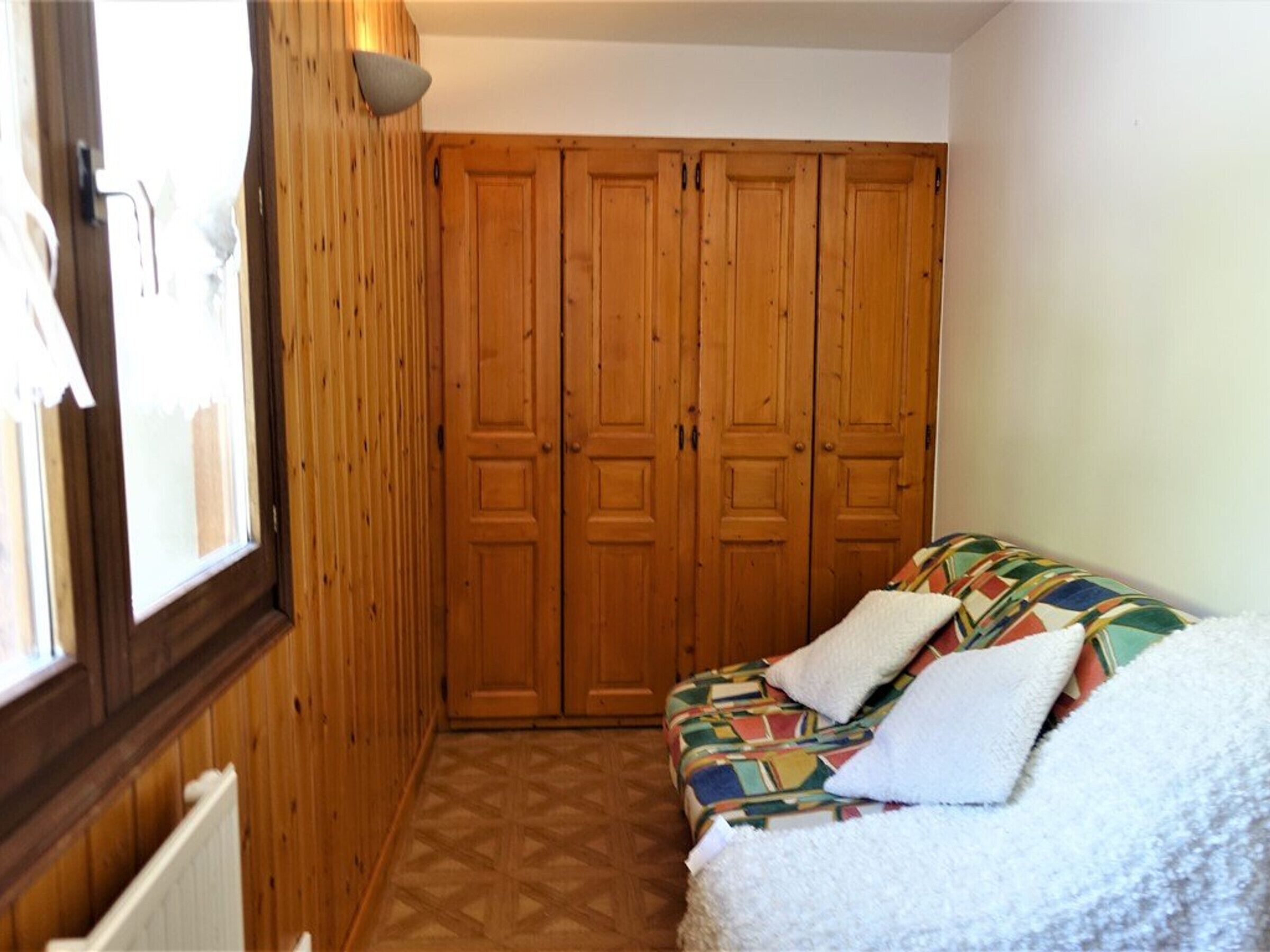 A room with a wooden wardrobe, a patterned sofa, and a window.
